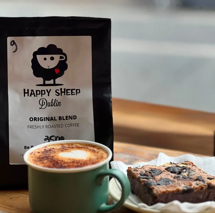 A bag of Happy Sheep Original Blend beside a coffee in a green mug, with a heart in the foam, and a chocolate brownie