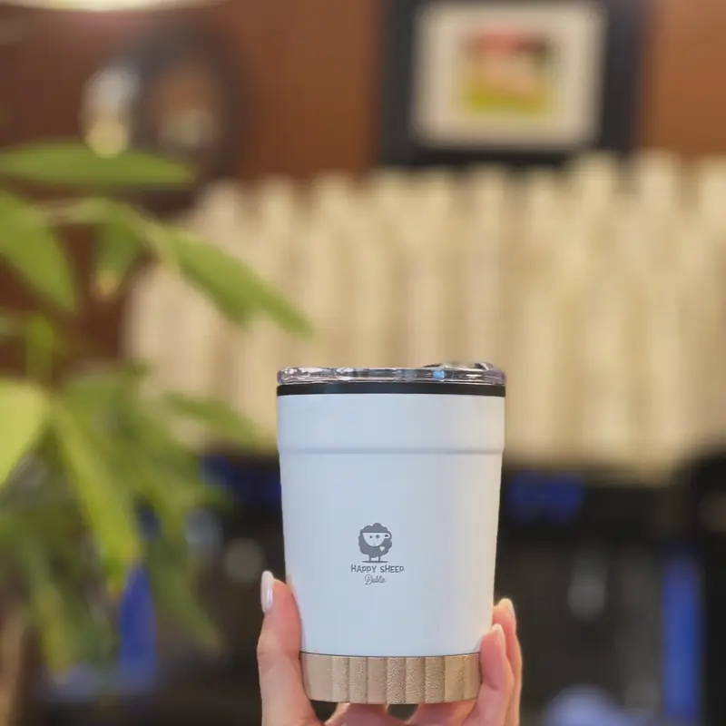 A hand holding a white insulated Happy Sheep Dublin cup with a wooden base