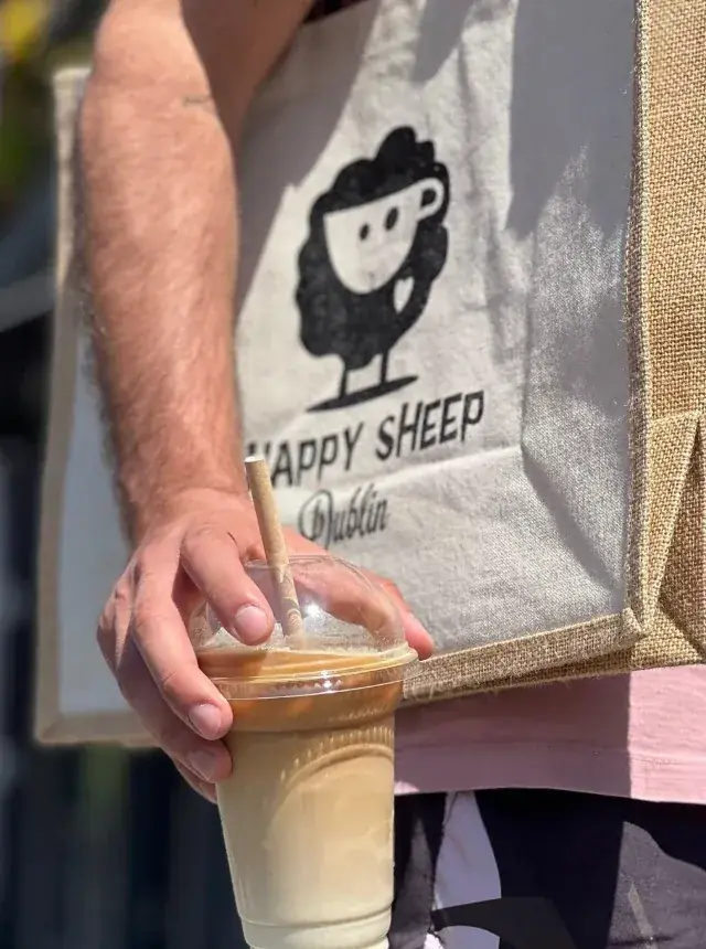 Iced latte with a paper straw, held in front of a Happy Sheep Dublin tote bag in the sun